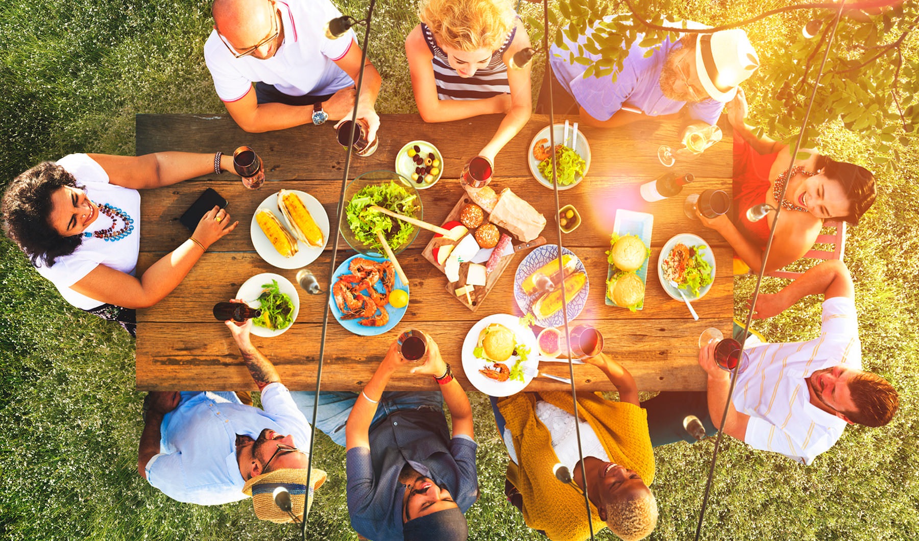 overhead view, looking down at a large picnic table full of food, surrounded by a group of people laughing and drinking 